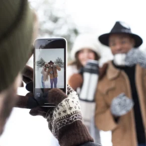 Vänner klädda i vinterkläder poserar för ett foto i snön medan en person fotograferar dem med mobilen.
